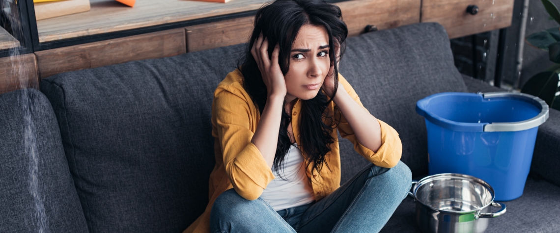 Frustrated brunette woman sitting on sofa under leaking ceiling