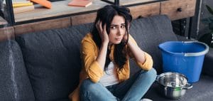 Frustrated brunette woman sitting on sofa under leaking ceiling
