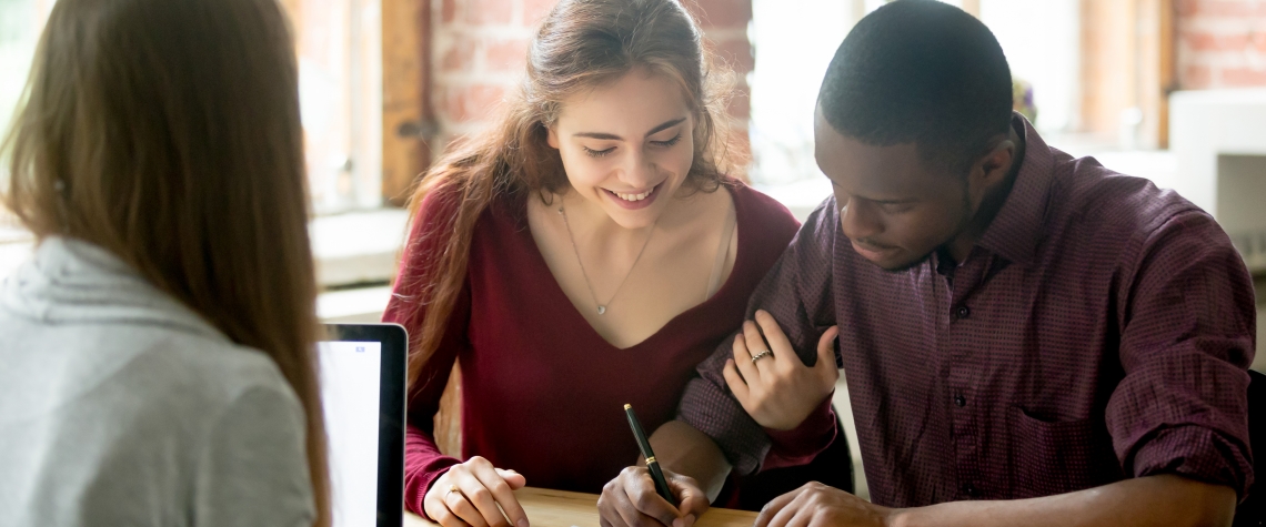 Smiling wife watching african american husband signing purchase deal from female real estate agent. Multiracial diverse couple buying first home together, making loan, legalizing mortgage.