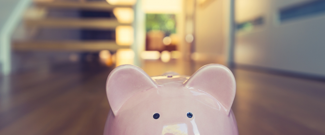 Saving for a home concept. Piggy bank standing at front door with house interior in the background. Pig is pink in foreground and house is modern style. Copy space.