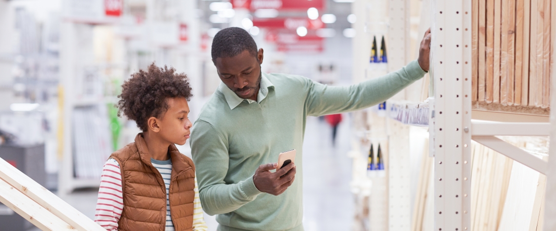 Waist up portrait of African-American father and son shopping together in hardware store and using smartphone while choosing wooden boards for home improvement