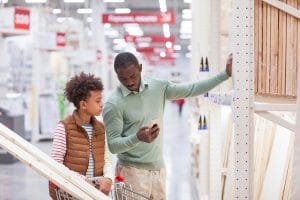 Waist up portrait of African-American father and son shopping together in hardware store and using smartphone while choosing wooden boards for home improvement