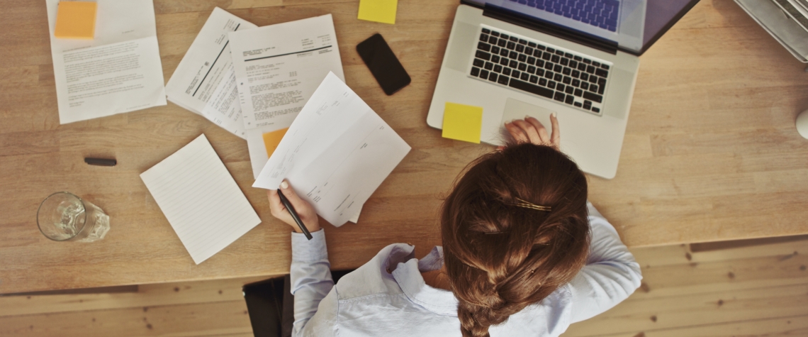 Businesswoman working at her office desk with documents and laptop High angle view of an young brunette working at her office desk with documents and laptop. Businesswoman working on paperwork.