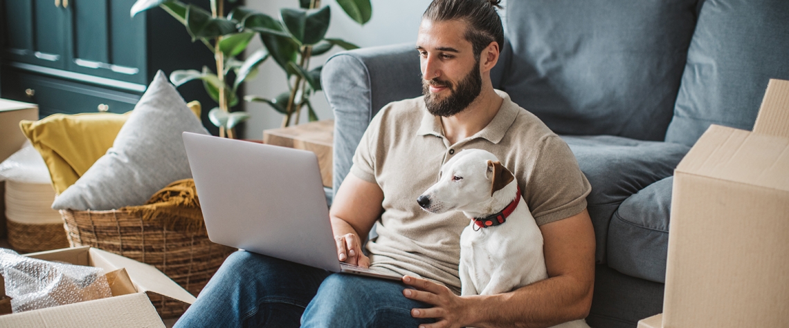 Young men moving in new house. He is happy and using laptop to organize everything. His pet dog is with him.