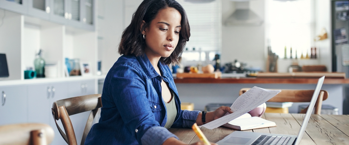 Shot of a young woman using a laptop and  going through paperwork while working from home