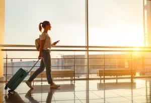 A young woman wearing white sneakers, blue jeans, a white t-shirt, and a small tan backpack, with her hair in a ponytail, holds her cell phone, passport, and plane ticket in one hand and pulls a teal rolling suitcase behind her with the other while walking through an airport bathed in golden light from the setting sun.