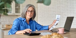 Adult senior 60s woman calculating paper documents at home at pc. Serious middle aged woman at table working on bank payments household online with computer technologies.