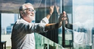 A man with white hair, white facial hair, dark glasses, and a white dress shirt smiles as he looks out over a sunny city through the large glass windows of an office building.