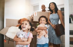 Image Alt Text A joyful family stands in their new home, surrounded by moving boxes. Two young children, holding stuffed animals, look excitedly around while their smiling parents stand in the background with a box and suitcase.