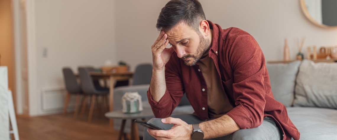 An anxious adult man reading bad news on his phone while sitting on a couch A worried adult man reading bad news and text messages on his phone while sitting on a couch in his apartment.