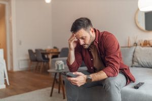 A worried adult man reading bad news and text messages on his phone while sitting on a couch in his apartment.