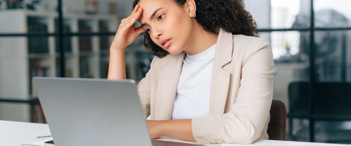 Upset frustrated latino or hispanic curly haired business woman, corporate financial manager, sits in a modern office, sadly looking at a laptop screen, reading bad news, received bad financial report