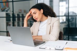 Upset frustrated latino or hispanic curly haired business woman, corporate financial manager, sits in a modern office, sadly looking at a laptop screen, reading bad news, received bad financial report